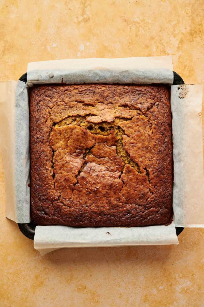 A square Pumpkin Cake baked in a parchment-lined pan on a beige surface, with visible cracks on the top.
