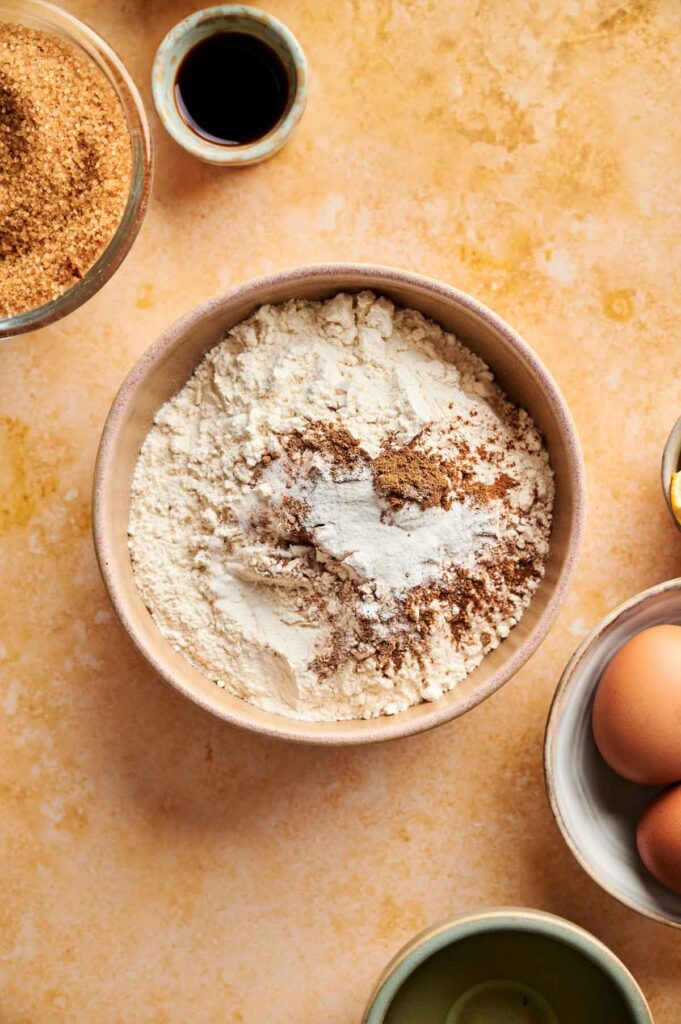 A bowl of flour mixed with brown sugar and spices sits on a countertop, surrounded by eggs, brown sugar, and a small bowl of liquid&mdash;ingredients ready to be transformed into a delicious pumpkin cake.