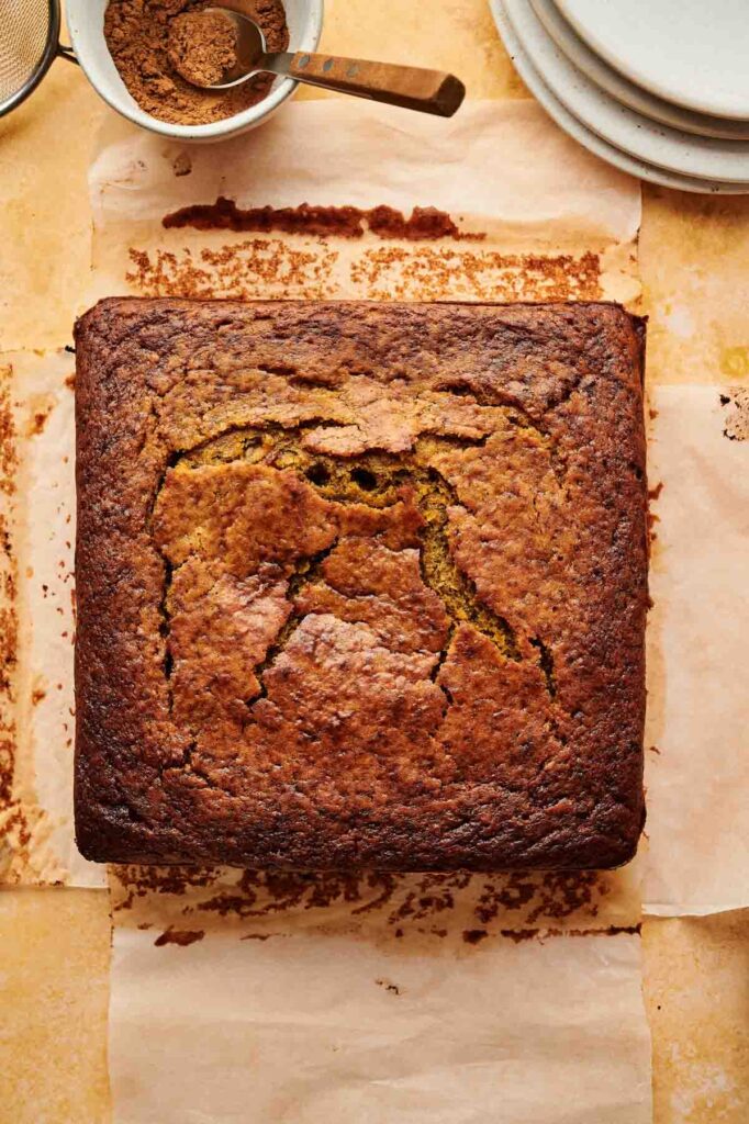 A square loaf of baked banana bread sits on parchment paper, its cracked surface resembling a rustic pumpkin cake. Plates and a bowl of baking ingredients are arranged nearby.