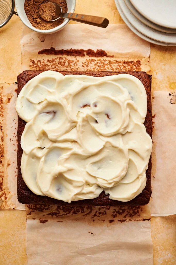 A square Pumpkin Cake with thick white frosting sits on parchment paper, with a dish of brown spice, a spoon, and stacked plates in the background.