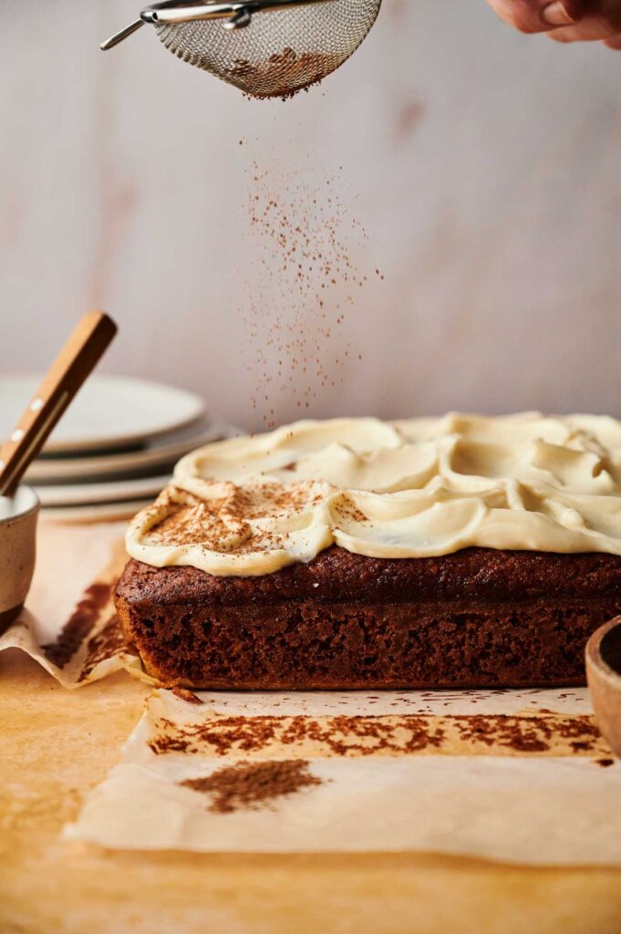 A hand sprinkles cocoa powder over a rectangular Pumpkin Cake with white frosting on parchment paper, with plates and utensils nearby.