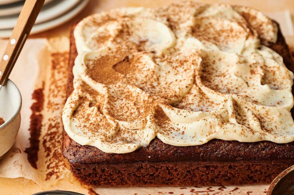 A square pumpkin cake with swirled cream cheese frosting and a dusting of cocoa powder on top, placed on parchment paper.