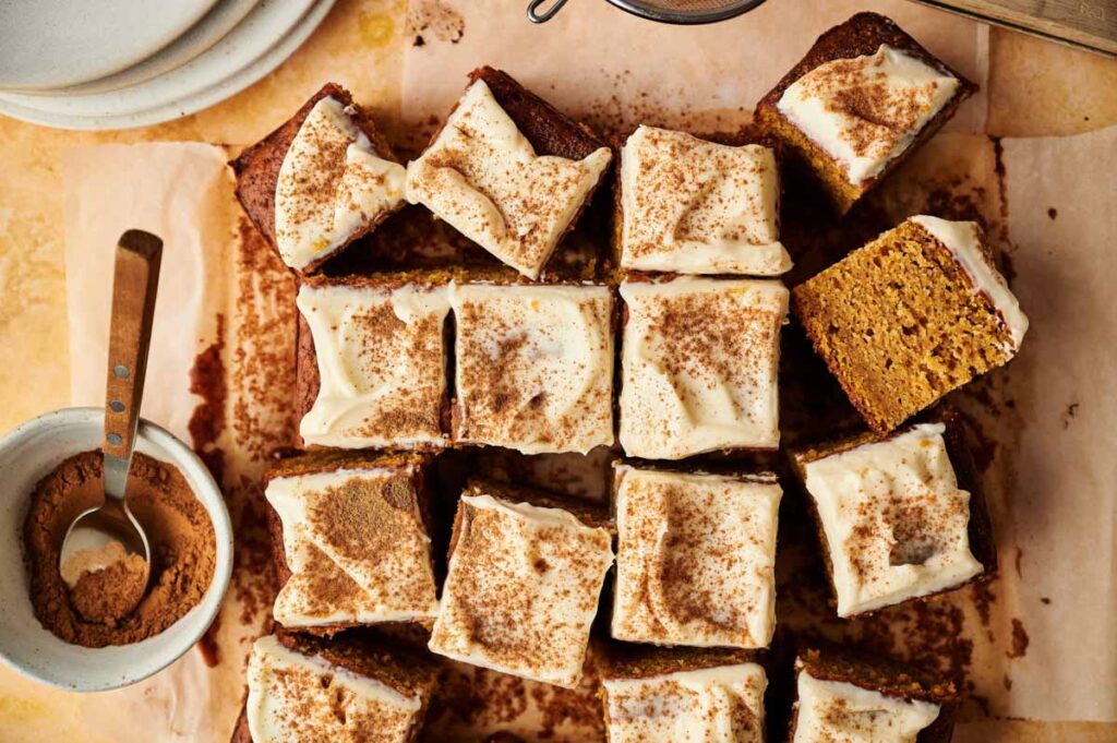 A batch of frosted Pumpkin Cake squares topped with cinnamon, arranged on parchment paper alongside a bowl of ground cinnamon and a spoon.