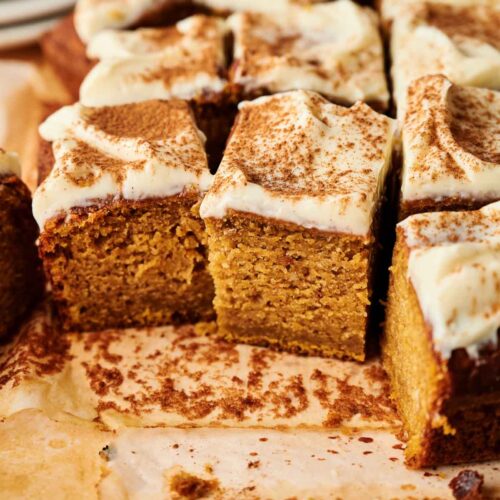 Square pieces of Pumpkin Cake, frosted and topped with a dusting of cinnamon, are arranged on a sheet of parchment paper, with a few slices cut and separated.
