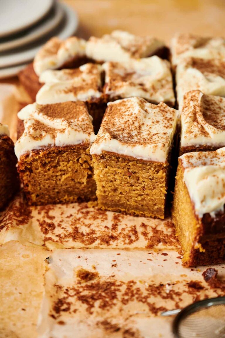 Square pieces of Pumpkin Cake, frosted and topped with a dusting of cinnamon, are arranged on a sheet of parchment paper, with a few slices cut and separated.