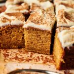 Close-up of delicious Pumpkin Cake squares topped with cream cheese frosting and a dusting of cinnamon, arranged on parchment paper.