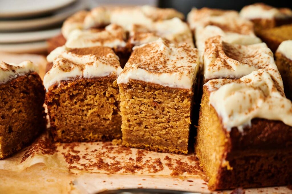 Close-up of Pumpkin Cake slices with cream cheese frosting and a dusting of cinnamon, arranged on parchment paper.