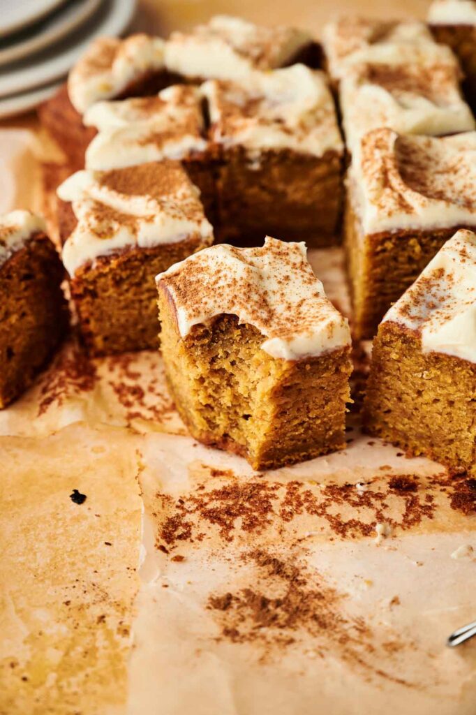 Squares of frosted Pumpkin Cake with cream cheese icing and a dusting of cinnamon are arranged on parchment paper, with one piece pulled slightly forward.