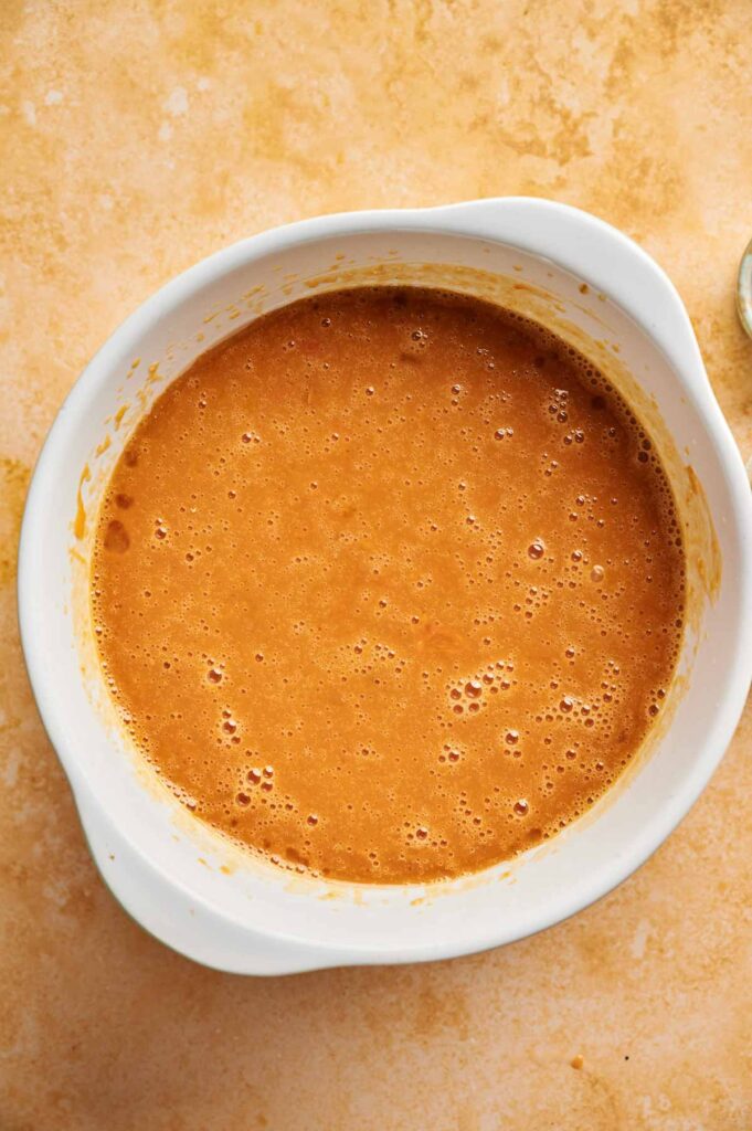 A white mixing bowl filled with light brown Pumpkin Cake batter sits on a tan surface.