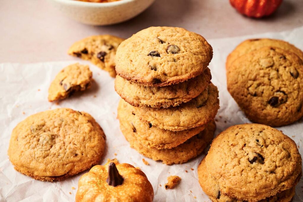 A stack of Pumpkin Chocolate Chip Cookies on parchment paper, with more cookies, a small decorative pumpkin, and a bowl in the background.