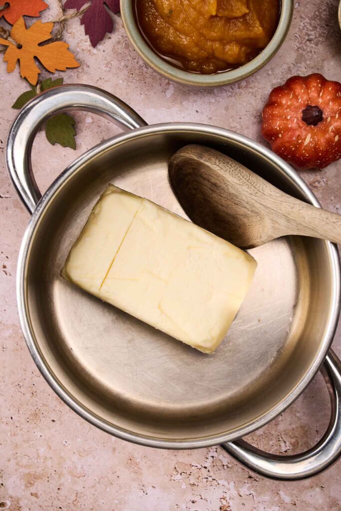 A stick of butter melts in a stainless steel pot with a wooden spoon, surrounded by autumn-themed decorations and a bowl of pumpkin puree&mdash;perfect for making Pumpkin Chocolate Chip Cookies.