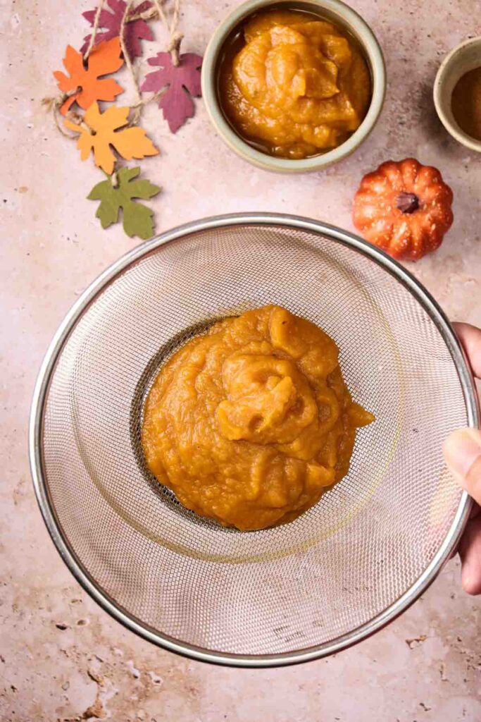 Pumpkin puree being strained through a metal sieve, with a bowl of pumpkin puree ready for baking Pumpkin Chocolate Chip Cookies, surrounded by decorative leaves and a small faux pumpkin on a light surface.