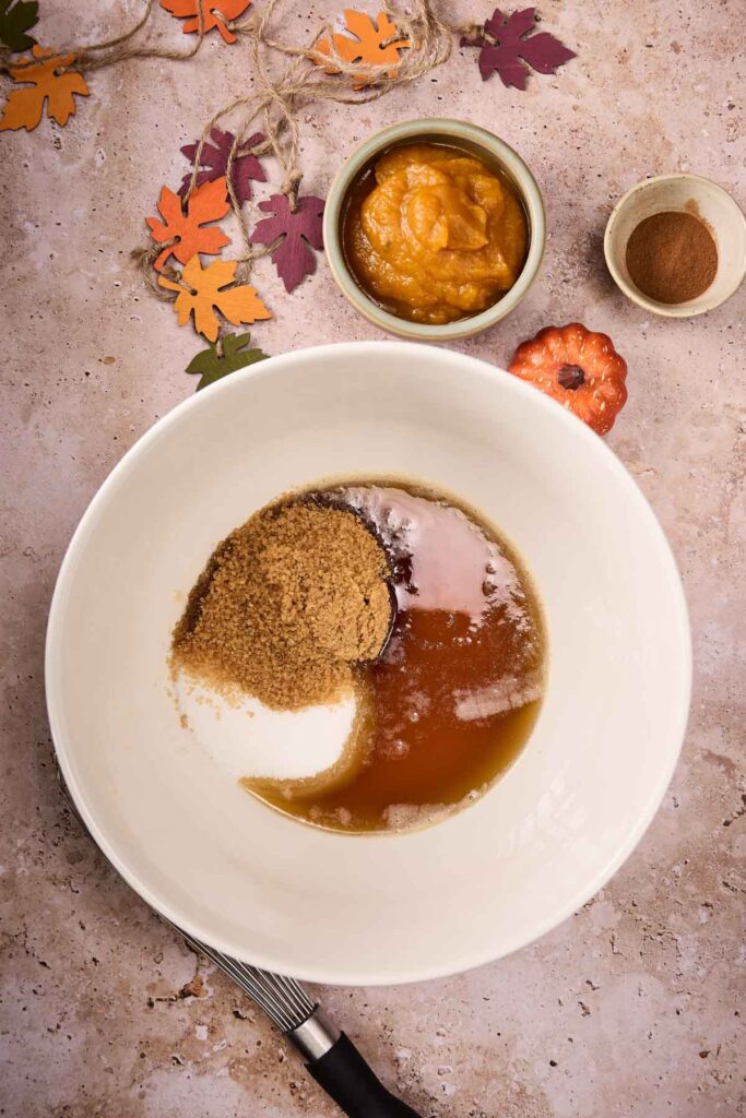 A white mixing bowl with brown sugar, white sugar, and a liquid ingredient&mdash;perfect for making Pumpkin Chocolate Chip Cookies. Nearby are bowls with pumpkin puree and spices, fall leaf decorations, and a whisk completing the cozy scene.