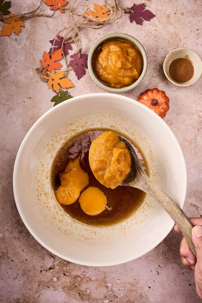 A hand holds a wooden spoon with pumpkin puree over a bowl of eggs and brown sugar, ready to mix up Pumpkin Chocolate Chip Cookies; small bowls of pumpkin puree and spices sit nearby.