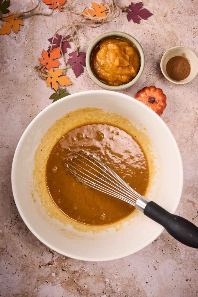 A mixing bowl with a whisk holds brown batter for Pumpkin Chocolate Chip Cookies. Nearby are a bowl of pumpkin puree, a bowl of spices, and autumn leaf decorations on a beige surface.