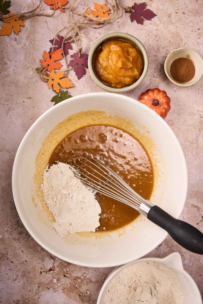 A mixing bowl with wet and dry ingredients being whisked for Pumpkin Chocolate Chip Cookies, surrounded by a bowl of pumpkin puree, a bowl of spices, and autumn leaf decorations.