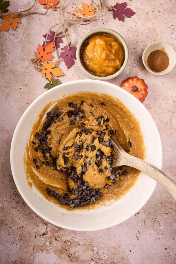 A mixing bowl with Pumpkin Chocolate Chip Cookies dough and a wooden spoon, surrounded by a bowl of pumpkin puree, a bowl of cinnamon, and autumn leaf decorations.