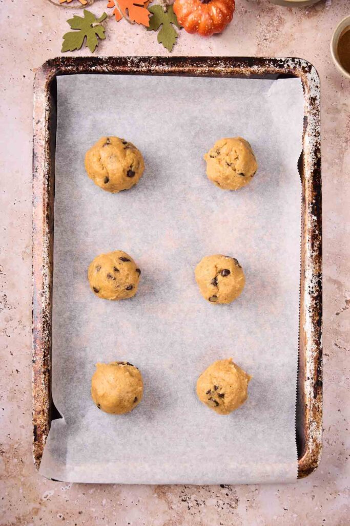 A baking sheet lined with parchment paper holds six evenly spaced raw Pumpkin Chocolate Chip Cookie dough balls, ready to be baked. Autumn-themed decorations are partially visible at the top.