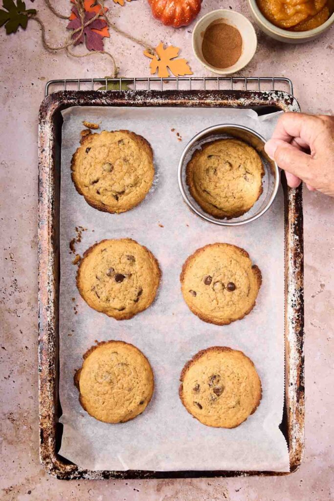 A baking sheet with six Pumpkin Chocolate Chip Cookies on parchment paper; a hand holds a round metal cutter over one cookie. Autumn leaves and spices are nearby.
