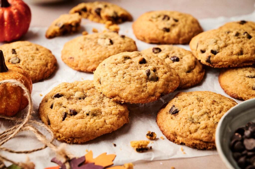A batch of Pumpkin Chocolate Chip Cookies on parchment paper, surrounded by a small pumpkin, twine, and a bowl of chocolate chips.