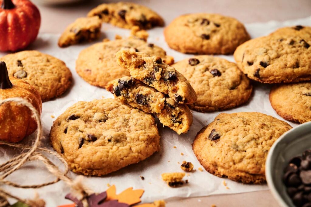 A batch of Pumpkin Chocolate Chip Cookies on parchment paper, with one cookie broken in half to reveal the gooey chocolate chips inside. Small pumpkins and autumn decorations are nearby.