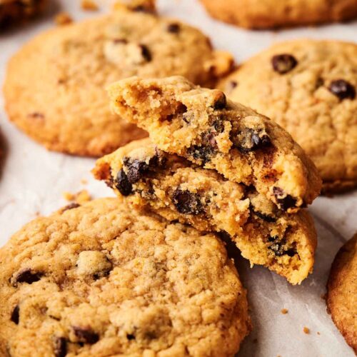 A close-up of several Pumpkin Chocolate Chip Cookies on parchment paper, with one cookie broken in half to reveal the soft, spiced interior and gooey chocolate chips.
