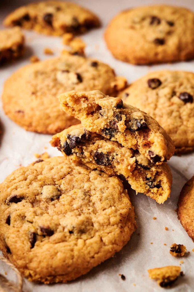 A close-up of several Pumpkin Chocolate Chip Cookies on parchment paper, with one cookie broken in half to reveal the soft, spiced interior and gooey chocolate chips.
