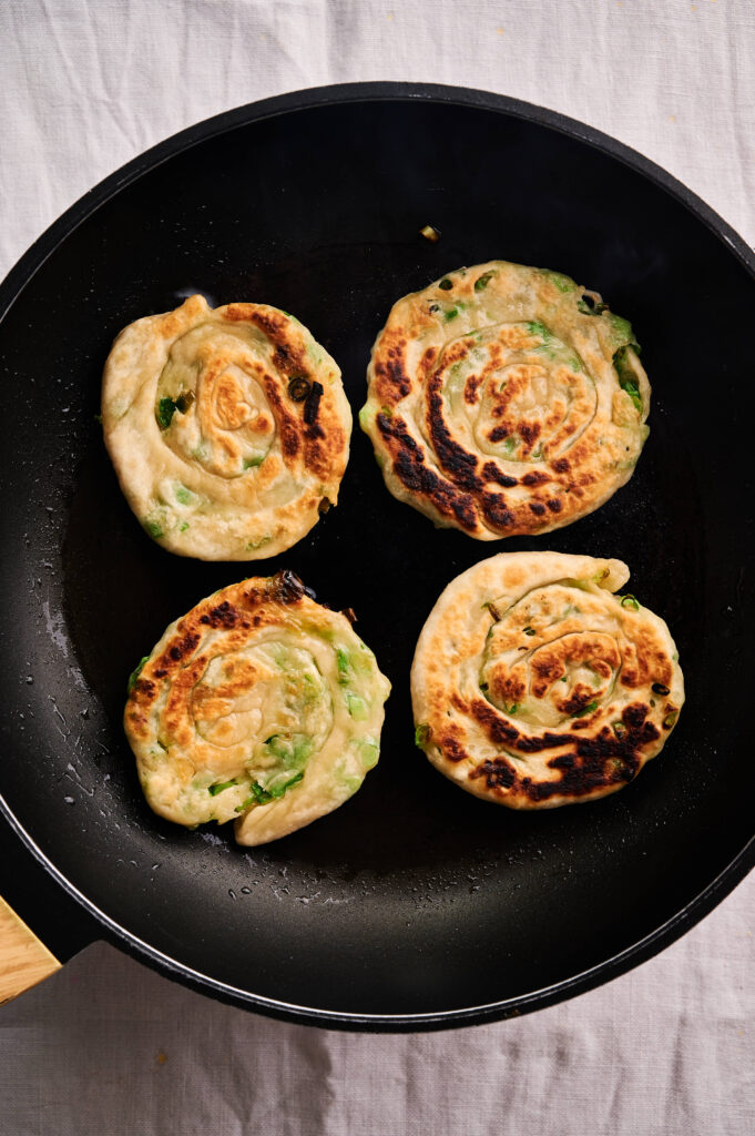 Four golden scallion pancakes are sizzling in a black skillet, set against a crisp white cloth background.