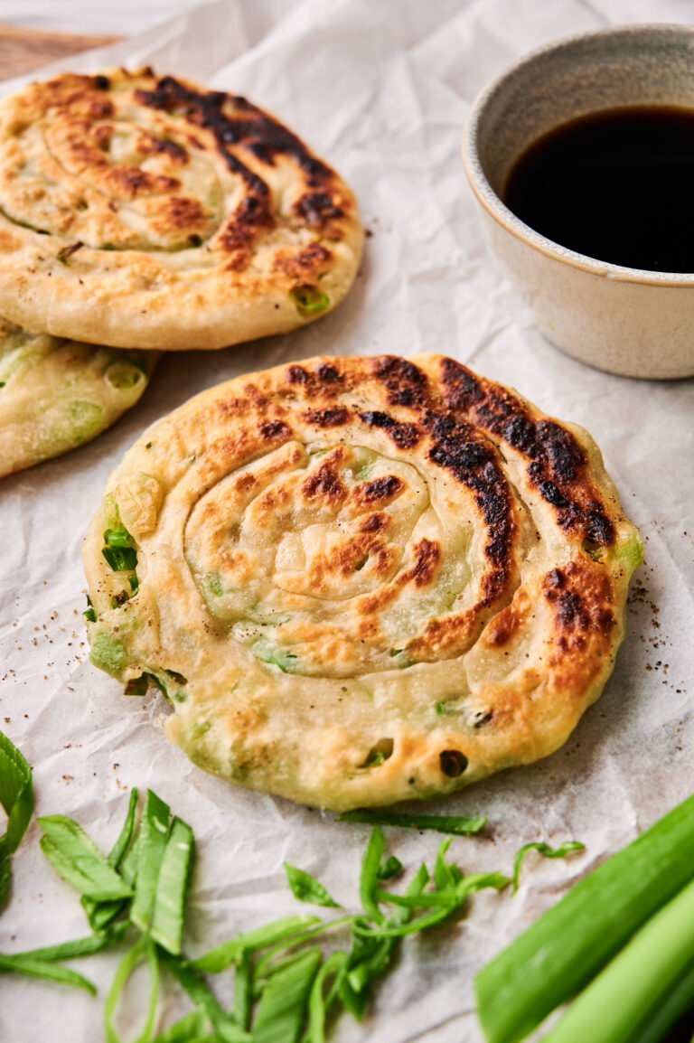 Two delicious Scallion Pancakes rest on parchment paper, garnished with sliced green onions and served next to a small bowl of dark dipping sauce.