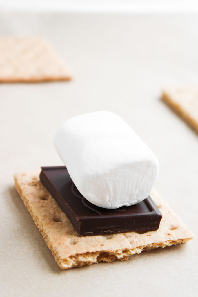 A large marshmallow and a square of chocolate sit on a graham cracker, evoking the delicious flavors of S&rsquo;mores Stuffed Cookies, with more graham crackers in the background.