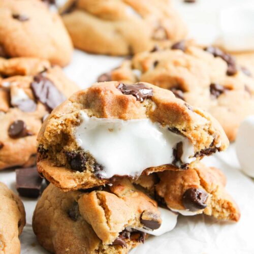 A close-up of S&rsquo;mores Stuffed Cookies with gooey marshmallow filling inside, surrounded by pieces of chocolate and graham crackers.