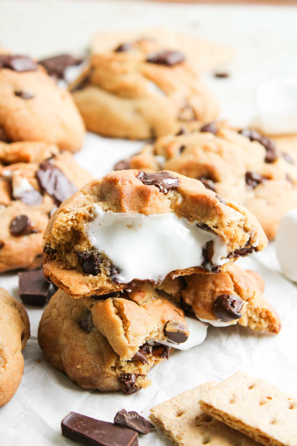 A close-up of S&rsquo;mores Stuffed Cookies with gooey marshmallow filling inside, surrounded by pieces of chocolate and graham crackers.