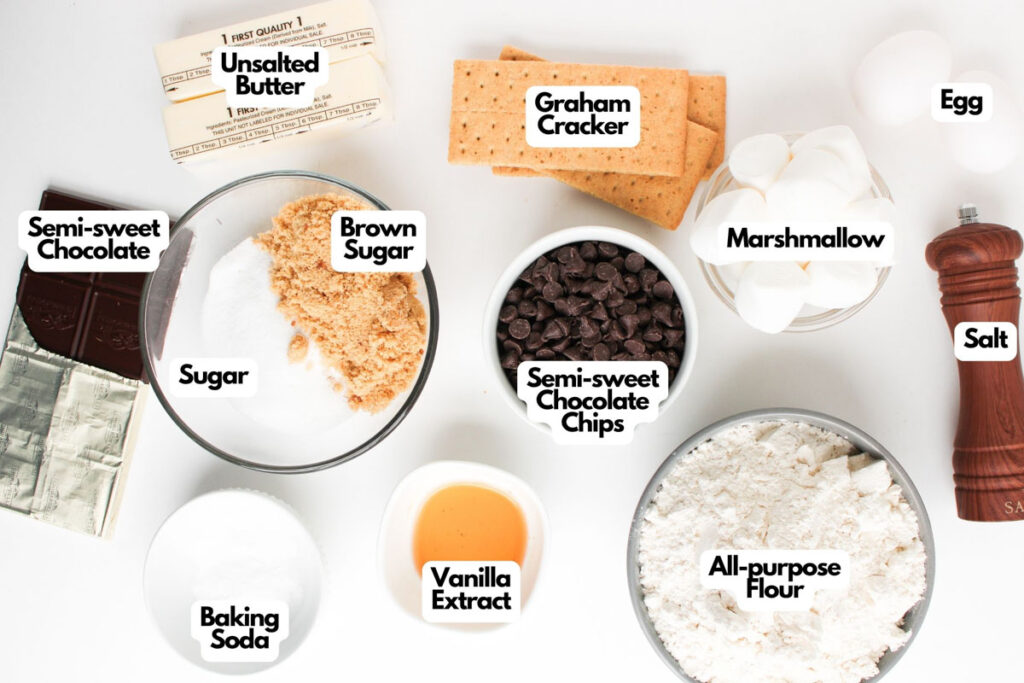 Top-down view of labeled baking ingredients for S&rsquo;mores Stuffed Cookies, including butter, sugars, chocolate, graham crackers, marshmallows, chocolate chips, flour, baking soda, vanilla extract, salt, and an egg.
