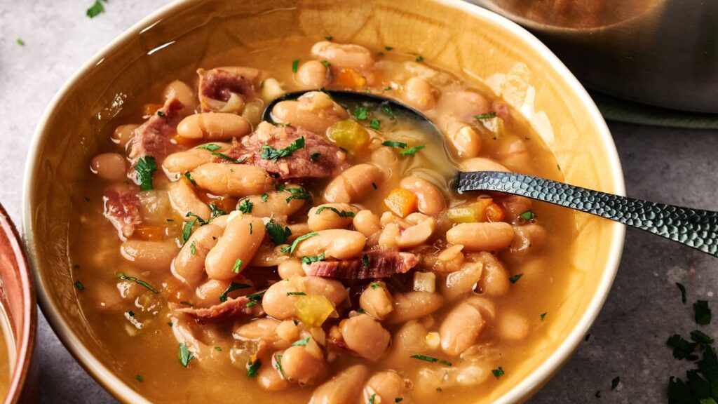 A bowl of bean and ham soup, garnished with chopped herbs, with a spoon resting inside the bowl.