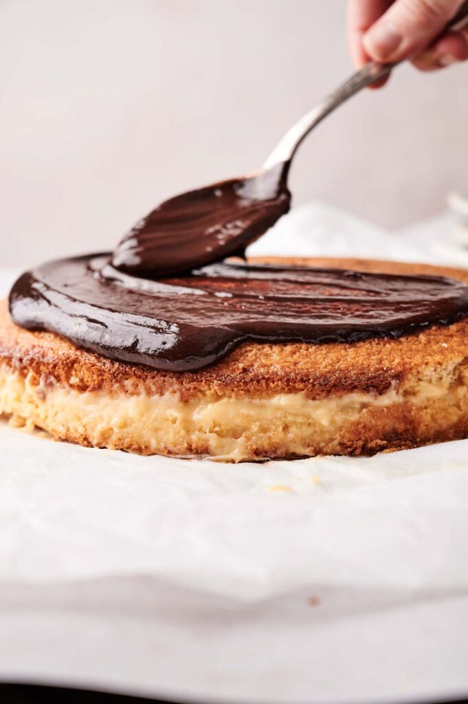 A hand spreads rich chocolate ganache over the top of a round, layered Boston Cream Pie on parchment paper.