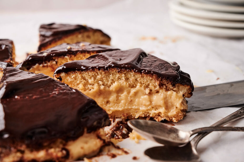 A slice of Boston Cream Pie with yellow sponge cake, creamy filling, and chocolate glaze is served with a cake server and two spoons on a white surface.