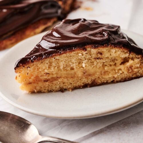 A slice of classic Boston Cream Pie with rich chocolate glaze and silky custard filling is served on a white plate next to a spoon.