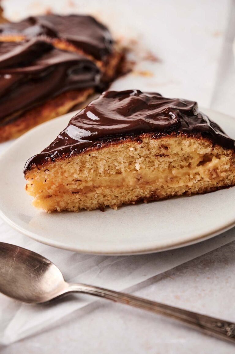 A slice of classic Boston Cream Pie with rich chocolate glaze and silky custard filling is served on a white plate next to a spoon.