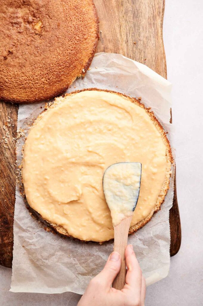 A hand spreads creamy frosting on a round Boston Cream Pie layer with a spatula; another unfrosted cake layer sits nearby on a wooden surface.