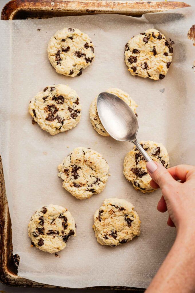A hand uses a spoon to shape unbaked Cannoli Cookies with chocolate chips on a parchment-lined baking sheet.