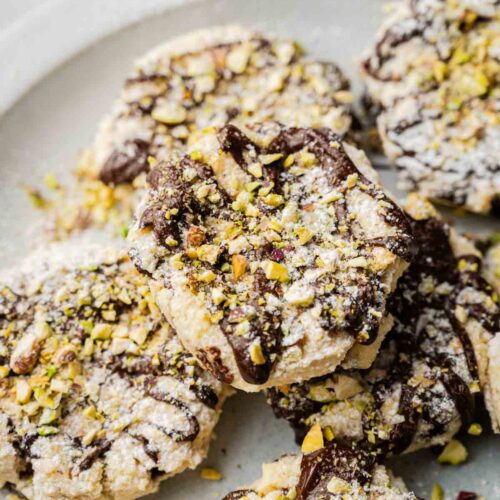 A plate of Cannoli Cookies topped with powdered sugar, drizzled with chocolate, and sprinkled with chopped pistachios.