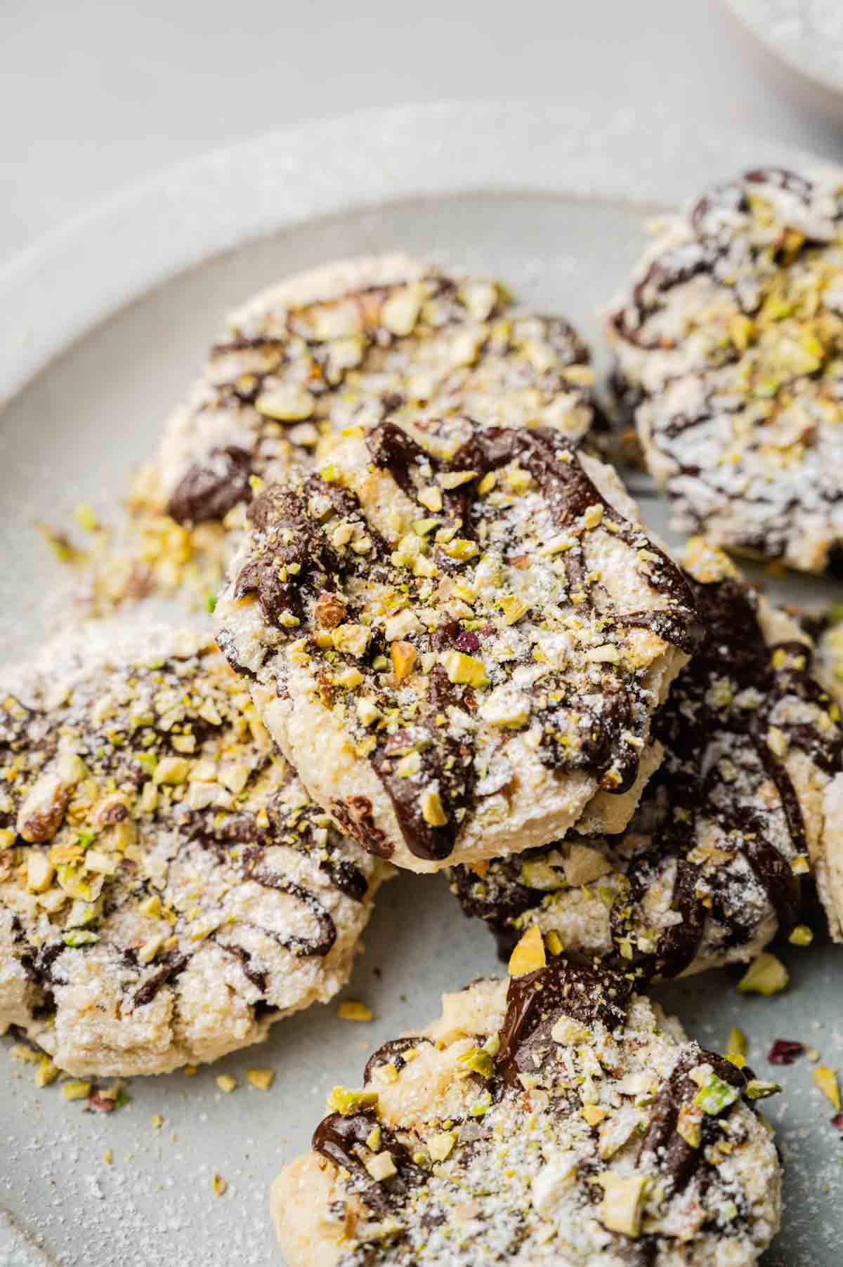 A plate of Cannoli Cookies topped with powdered sugar, drizzled with chocolate, and sprinkled with chopped pistachios.