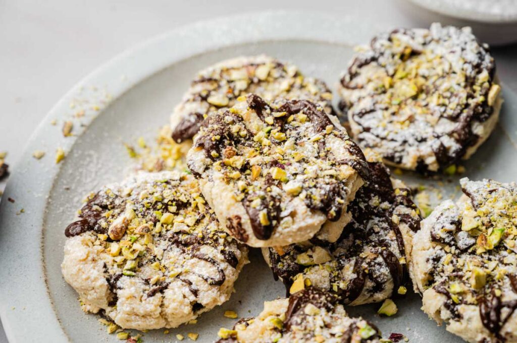 A plate of Cannoli Cookies topped with powdered sugar, chocolate drizzle, and chopped pistachios.