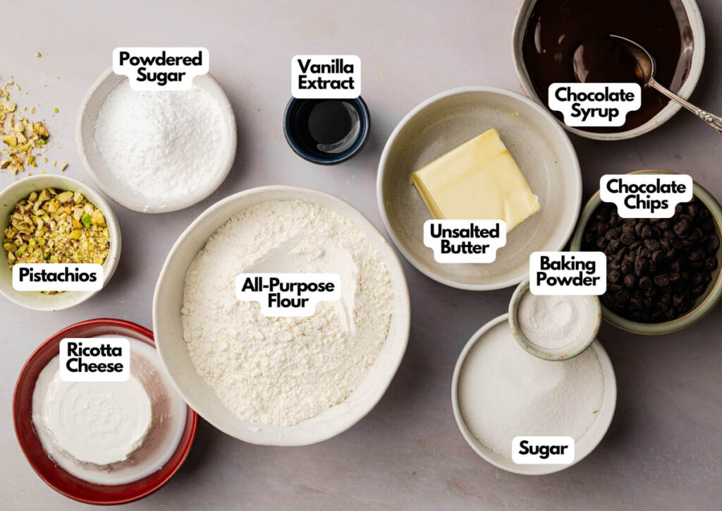 Bowls containing ingredients for baking Cannoli Cookies, including pistachios, ricotta cheese, powdered sugar, all-purpose flour, vanilla extract, unsalted butter, chocolate syrup, chocolate chips, baking powder, and sugar.