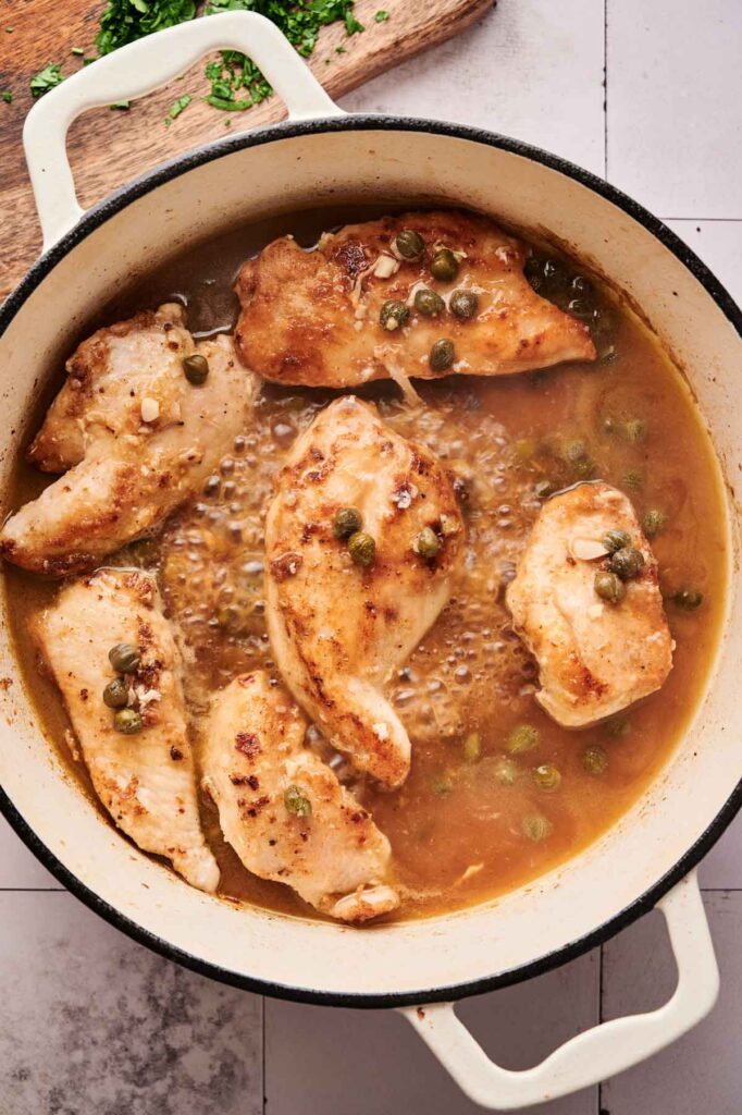 Six cooked chicken breasts in a light brown Chicken Piccata sauce with capers and garlic, served in a large white enameled pot on a tiled surface.