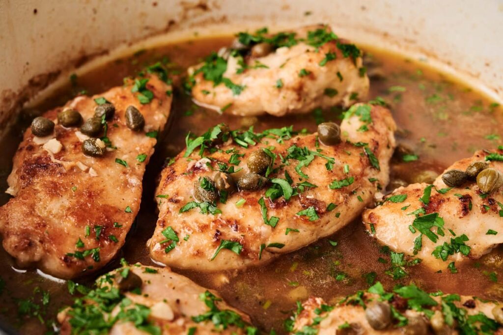 Chicken Piccata—tender chicken breasts cooked in a pan with a tangy brown sauce, garnished with chopped parsley and capers.