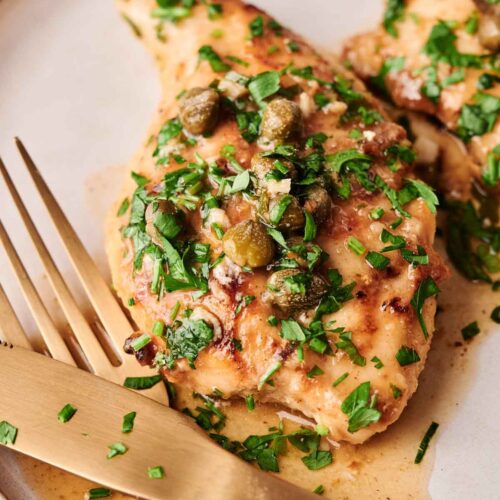 A classic Chicken Piccata: cooked chicken breast topped with capers and chopped parsley, served on a plate with a gold-colored fork and knife.