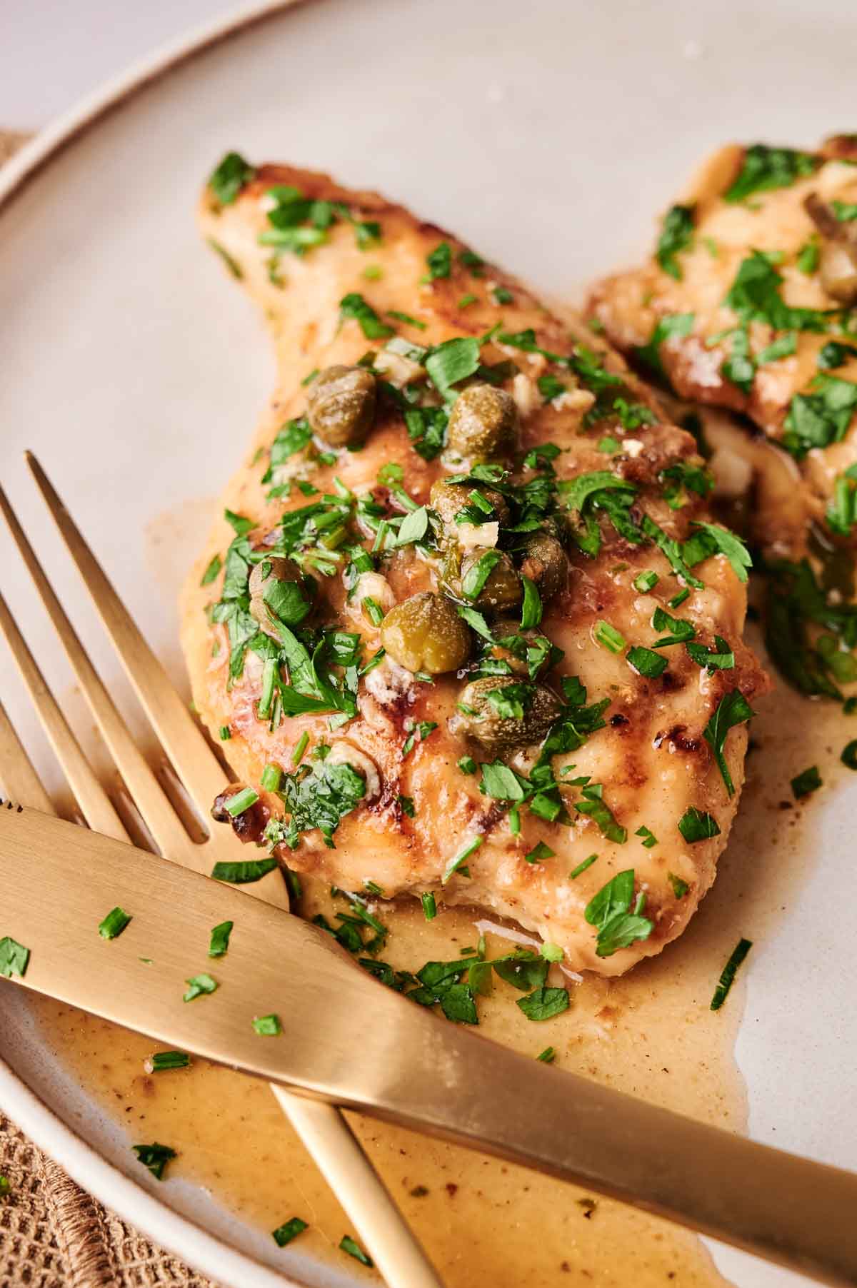 A classic Chicken Piccata: cooked chicken breast topped with capers and chopped parsley, served on a plate with a gold-colored fork and knife.