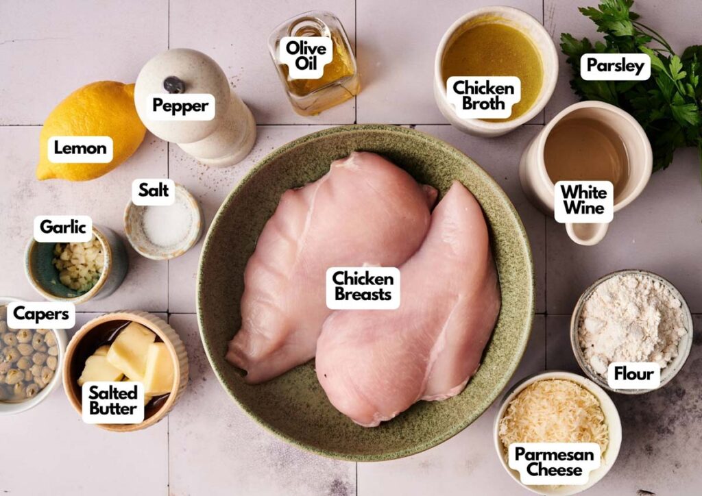 A bowl with raw chicken breasts surrounded by labeled ingredients for Chicken Piccata: olive oil, chicken broth, parsley, white wine, flour, parmesan cheese, salted butter, capers, garlic, lemon, salt, and pepper.