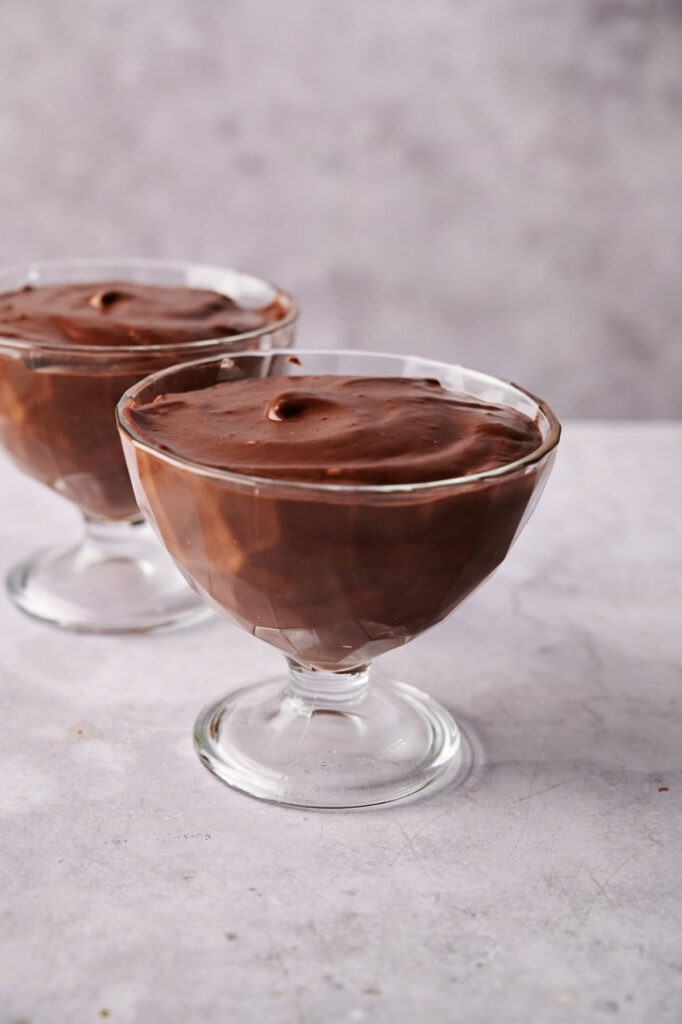 Two glass dessert cups filled with rich chocolate mousse, reminiscent of classic chocolate pudding, sit on a light gray surface with a plain background.